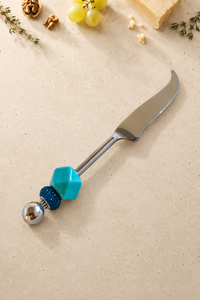 The Ananda decorative cheese knife with a silver-coloured curved blade, a geometric turquoise bead, a textured dark blue accent bead and a polished silver end bead.
