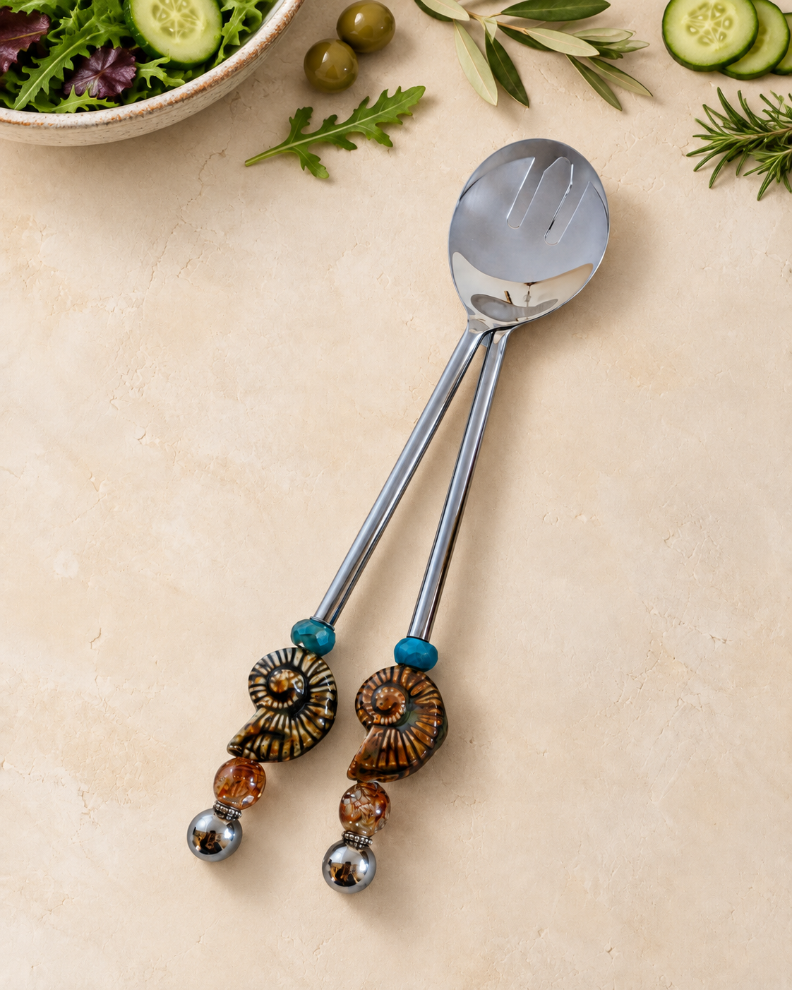 The Hanai decorative salad serving set with a silver-coloured spoon and slotted server, blue accent beads, spiral ammonite details and amber-coloured glass beads.