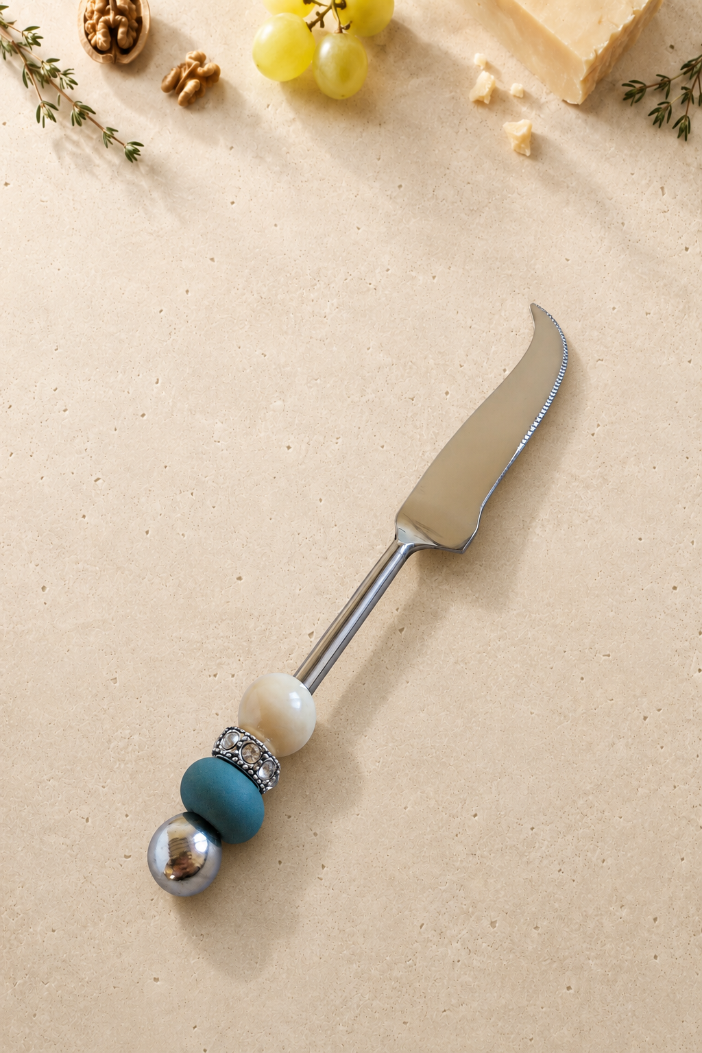 The Kasra cheese knife featuring a silver-coloured curved blade, an ivory bead, a matte blue accent bead and a polished silver end bead.