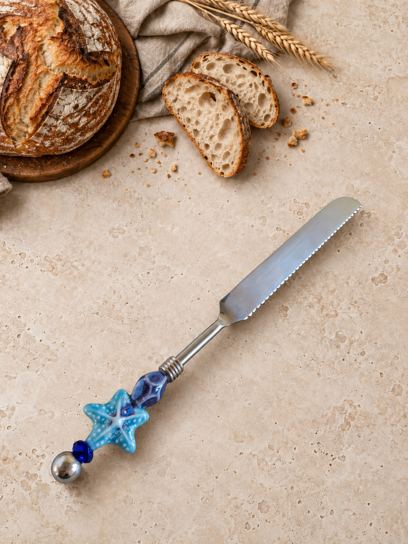 The Xavon handmade bread knife with a blue starfish beaded handle, styled on a light stone surface with rustic bread, sliced sourdough and wheat accents.